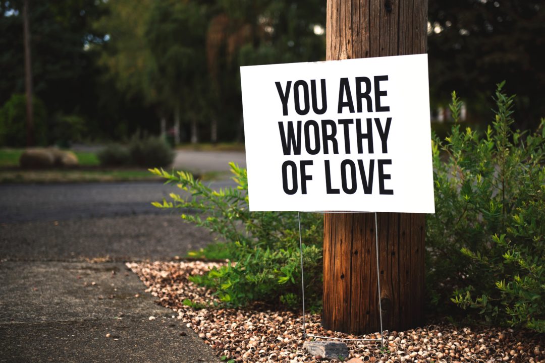 A white sign with black letters stating "You are worthy of love" posted on a tree in a suburban neighborhood.