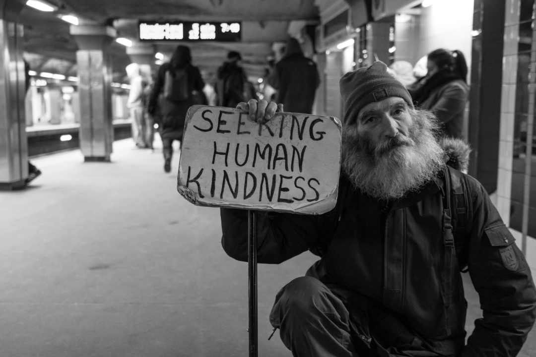 A homeless man holding up a sign that says "Seeking human kindness" in a handmade sign with black lettering.