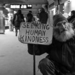 A homeless man holding up a sign that says "Seeking human kindness" in a handmade sign with black lettering.