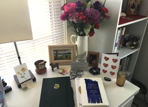 Flowers, painting, journal, books, and altar on white writing desk.